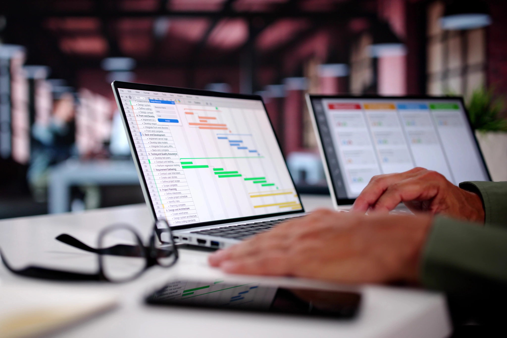 Laptop displaying a Gantt chart while someone types, with a Kanban board on a second screen.