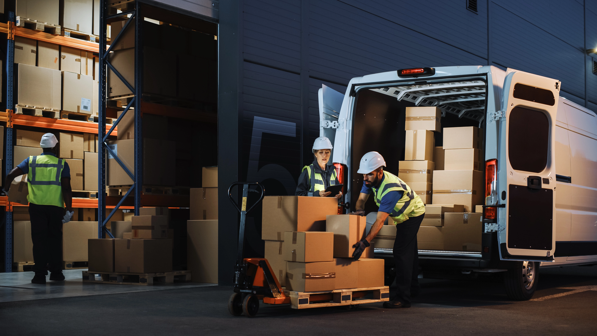 Warehouse team in hi-vis loading boxes onto a pallet and into an open delivery van at a loading bay.